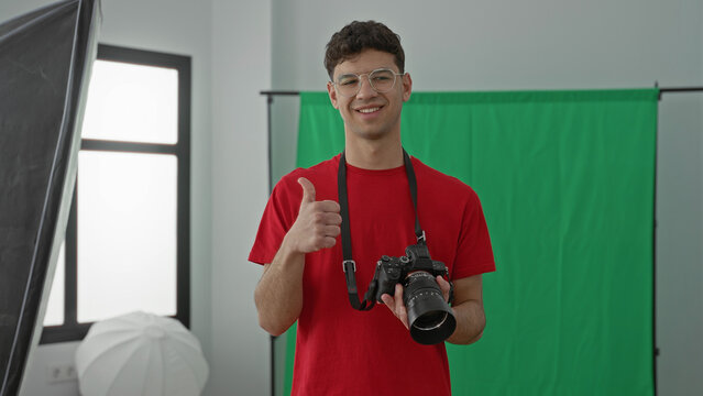 Young man photographer holding camera and giving thumbs up in studio with green screen and softbox lights, red shirt and glasses; creative confidence.