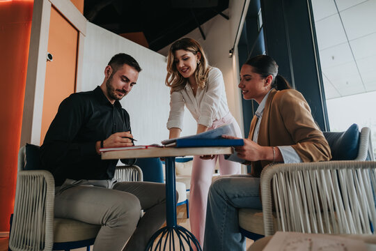 Three colleagues review documents and sign papers while discussing work. A casual remote work meeting shows collaboration and planning around a small table.