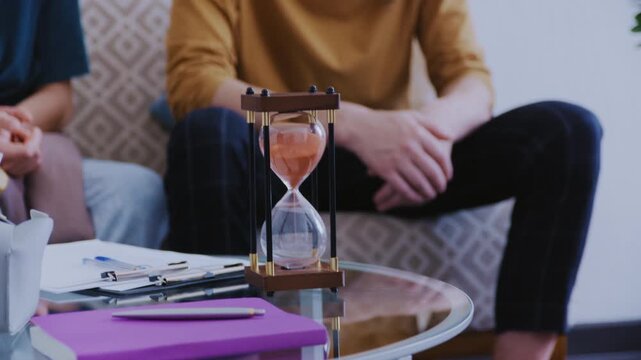 A professional resets a sand timer during a relationship support meeting while a couple discusses personal matters