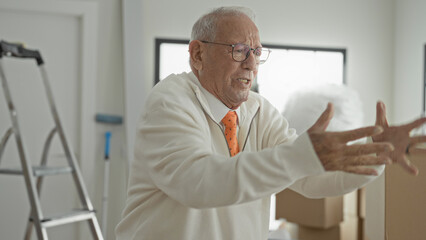 Fototapeta premium Elderly man, grey-haired and wearing glasses and an orange tie, gestures with hands while tossing packing foam amid moving boxes and a ladder inside a building; frustration.