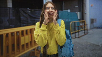 Fototapeta premium Teenage girl with backpack cupping hands at mouth calling on street near construction barriers, yellow sweater and visible braces; joyful energy.