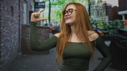 Fototapeta premium Redhead woman pinches fingers to indicate small size with her hand on a street, smiling broadly in glasses and green shirt; playful confidence.