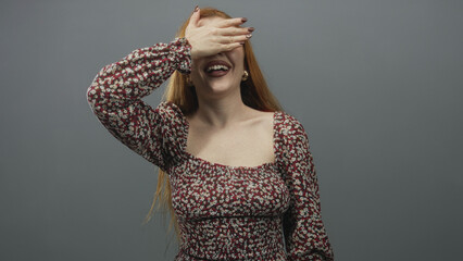 Woman covering her eyes with hand while smiling in a studio, young redhead in a floral dress showing shoulder and hoop earrings  playful joy. © Krakenimages.com