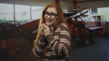 Fototapeta premium Redhead woman clasping hands and smiling with glasses, showing hands and teeth, posed by red farm machinery inside a building; contentment warmth.