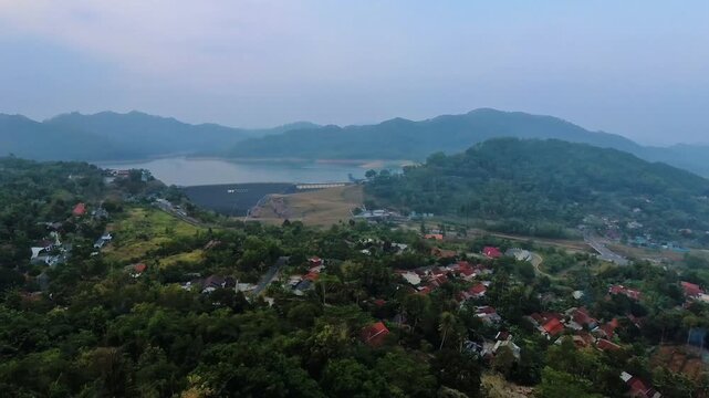 A high-angle panning shot showcasing a serene lake or reservoir nestled among lush tropical green hills and a small rural village under a misty morning sky. Ideal for travel promotions, environmental