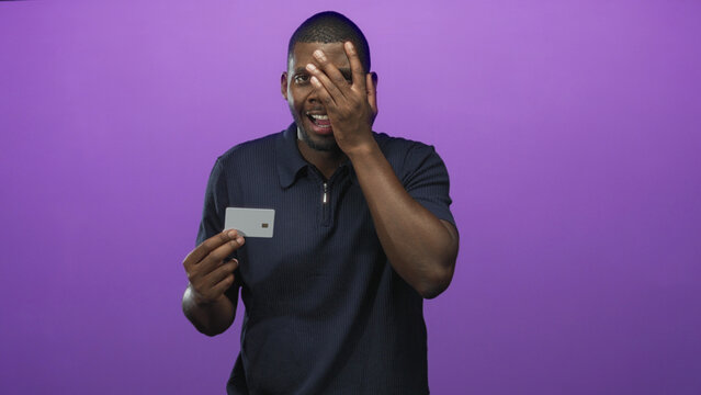Man holding creditcard and covering one eye with hand in a purple studio backdrop, mouth open in a startled expression; financial anxiety.