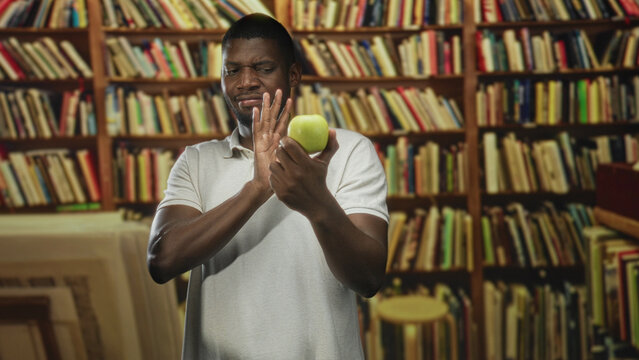 Man holding green apple and raising hand to shield face in library building; hesitation rejection disgust.