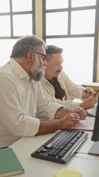 Two men coworkers at office desk, one holds smartphone, the other points pen to printed document with hands near keyboard in building; focused mentoring.
