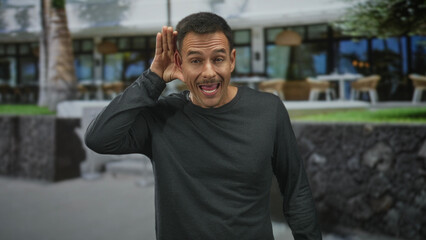 Fototapeta premium Man with moustache cups right hand to ear and tilts head while listening in front of a building cafe; curiosity engagement.