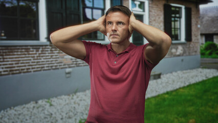 Fototapeta premium Man in red polo pressing hands to ears against a brick building facade, grimacing and squinting with visible forearms and hands; frustration reflection.