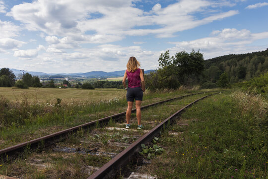 Back view of a woman standing on old railway tracks looking at a beautiful countryside landscape under blue sky. Summer travel and freedom concept