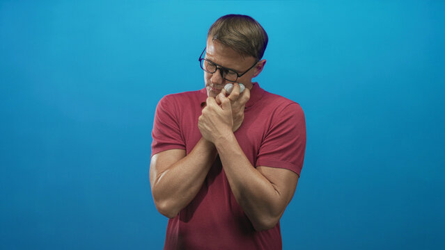 Man holding a contact lens case to his cheek while squinting and inspecting the small container in a studio with a blue backdrop; concentration.
