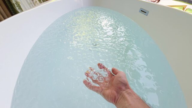 Hand Holds Clear Ice Cubes Above Cold Water in a Bathtub, Preparing an Ice Bath for Cryotherapy, Physical Recovery and Wellness