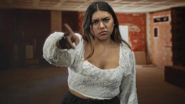 Woman hispanic plus size points finger at camera and touches temple with finger in unfinished building interior wearing white blouse; frustration defiance.