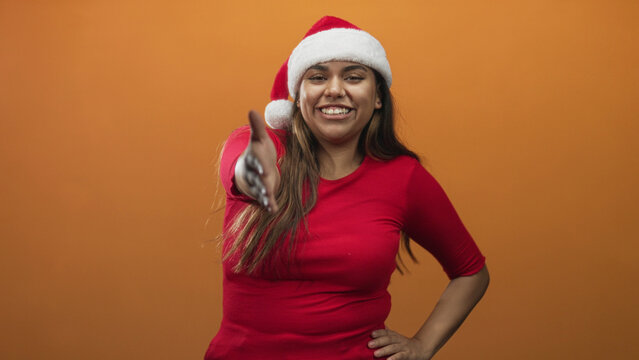 Woman extends right hand for handshake in studio wearing santa hat and red top while smiling and offering a greeting; playful welcome.