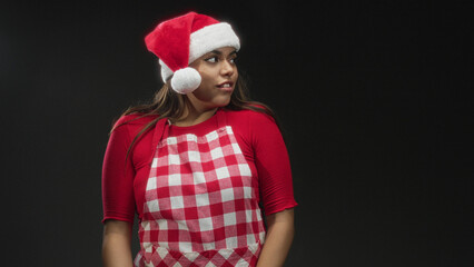 Fototapeta premium Woman in santa hat and red check apron, eyes closed and hands clasped in studio; holiday cheer contentment.