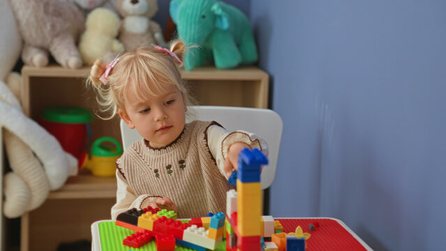 Little blonde girl building a colorful block tower while seated at a playroom table with stuffed plush toys on a shelf nearby; playful curiosity.