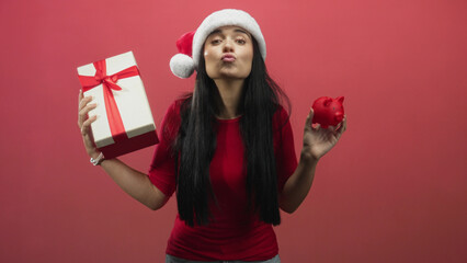 Fototapeta premium Woman wearing santa hat holding red piggy bank and wrapped gift box, smiling while presenting in studio; holiday saving joy.