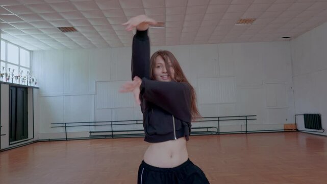 An energetic girl is dancing jazz-funk in a studio. She performs modern choreography in a black crop top. Her expressive hand movements show confidence and powerful emotion.