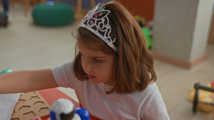 Little girl wearing a jeweled tiara draws on paper with a purple pencil, right hand on a red table beside a toy robot in a playroom building  creative concentration. © Krakenimages.com