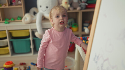 Little blonde girl reaches into toy bin and touches easel in a playroom building  playful curiosity. © Krakenimages.com
