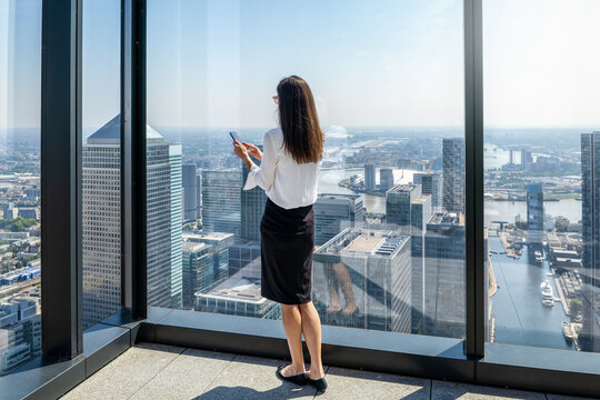 A business woman looking at her mobile phone in front of large floor to ceiling office windows with the urban skyline of London in the background