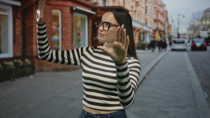Fototapeta premium Woman with palms out and hands raised in a stopping gesture on a city street storefront sidewalk wearing a striped top and glasses; personal boundary rejection.