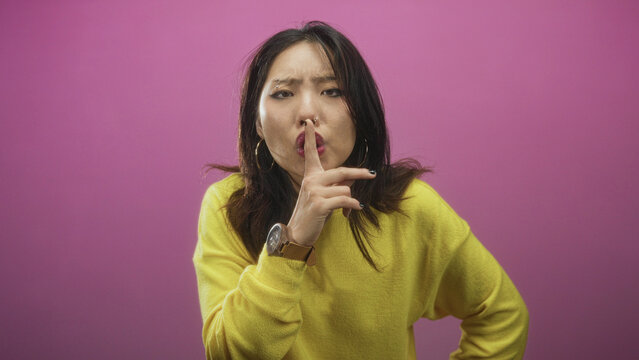 Woman in yellow sweater holds finger to lips shushing in studio against pink wall; secretive hush silence.