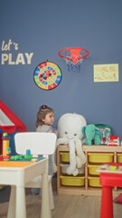 Girl holds ball and peeks behind toy shelf in colorful playroom with stuffed octopus, green elephant plush, block table and wall hoop  playful joy. © Krakenimages.com