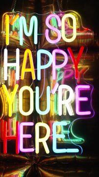 Colourful neon letters spelling out the positive message &lsquo;I&rsquo;m so happy you are here&rsquo;, surrounded by metallic balloons. A concept designed for a pleasant time. Party theme, positivity.