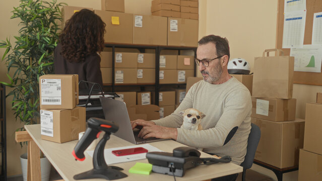 Man typing on laptop with small dog on lap and stacked shipping parcels, scanning labels with barcode reader in building; focused productivity.