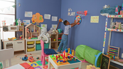African american boy reaching for wall dartboard while holding toy car in colorful classroom  playful curiosity. © Krakenimages.com