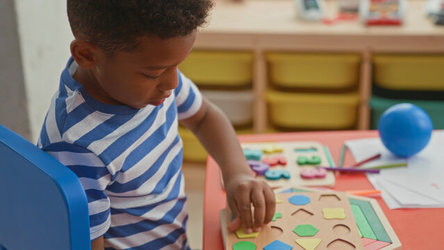 Boy placing a wooden shape puzzle piece with hands at a low table in a classroom inside a building, toys and papers nearby; concentration.