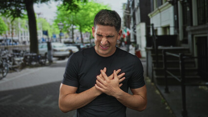 Fototapeta premium Young hispanic man grimacing clutching chest with bare hands on city street near bicycles and parked cars; heartattack emergency pain.