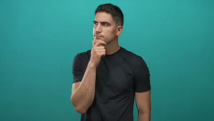 Fototapeta premium Man with hand on chin in studio with teal backdrop, wearing black tshirt and looking to the side; thoughtful reflection.