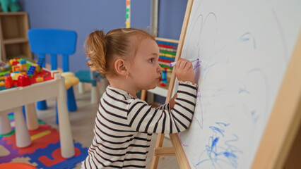 Little blonde girl drawing on a whiteboard with a purple marker at a wooden easel in a daycare building, small hand on board and intent expression  curiosity learning play. © Krakenimages.com