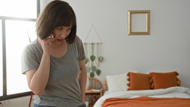 Woman holding smartphone to ear by bed and window, talking on phone inside a residential building; private thoughtful.