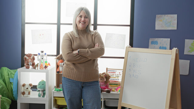 Woman teacher smiling with arms crossed beside whiteboard and easel in a classroom building with toys, abacus and wall drawings; confidence.