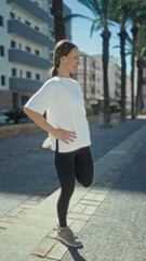 Fototapeta premium Woman stretching leg while balancing on sneaker, wearing sportswear and smartwatch on a sunny street with palm trees and buildings; steady determination.