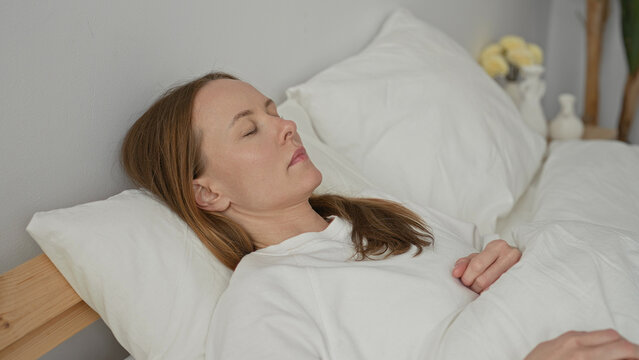 Woman sleeping with arm over head on pillow in a bedroom, bare forearm visible with hand resting on white duvet and wooden headboard visible; rest recovery serenity.