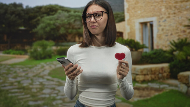 Woman holding smartphone and small red heart prop, frowning and squinting at messages while standing in front of a stone building courtyard; dating frustration.