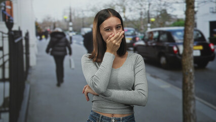 Fototapeta premium Young hispanic woman covering mouth with hand and slight smile on street, gray top and jeans visible; shy amusement.
