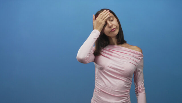 Woman with hand on forehead and eyes closed, slight wince and visible forehead skin, wearing pink off shoulder top in studio; fatigue reflection.