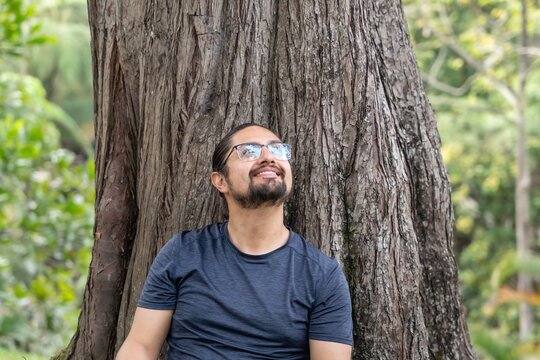 Smiling man leaning on large tree trunk looking up in park