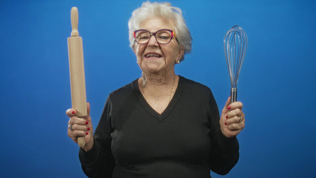 Grandmother holding a wooden rolling pin and metal whisk with red nails, smiling and gesturing with hands in a studio portrait; nostalgia joy.