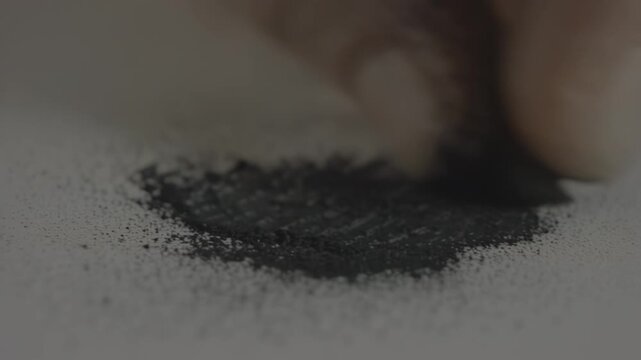 Close-up of hand pressing black powder onto white surface, gradually forming a distinct pile, showcasing the meticulous process of creating a textured design