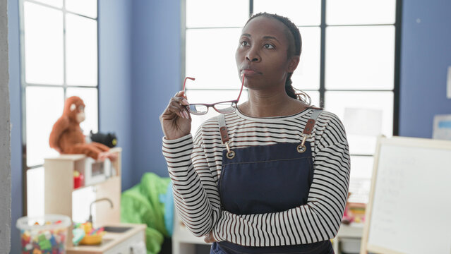 Woman holding glasses with hand near mouth in classroom building wearing apron and standing by whiteboard; pensive reflection.