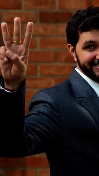 Showing numbers using fingers with confident expression, Indian Asian bearded ceo businessman in formal corporate attire demonstrating finger counting while standing in cozy office against brick wall