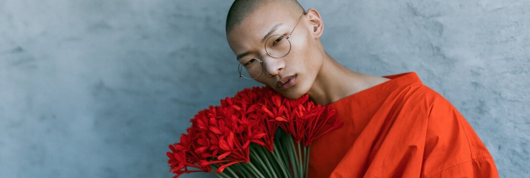 Contemporary portrait of androgynous model holding red gerbera flowers in orange robe for fashion editorial background and banner design