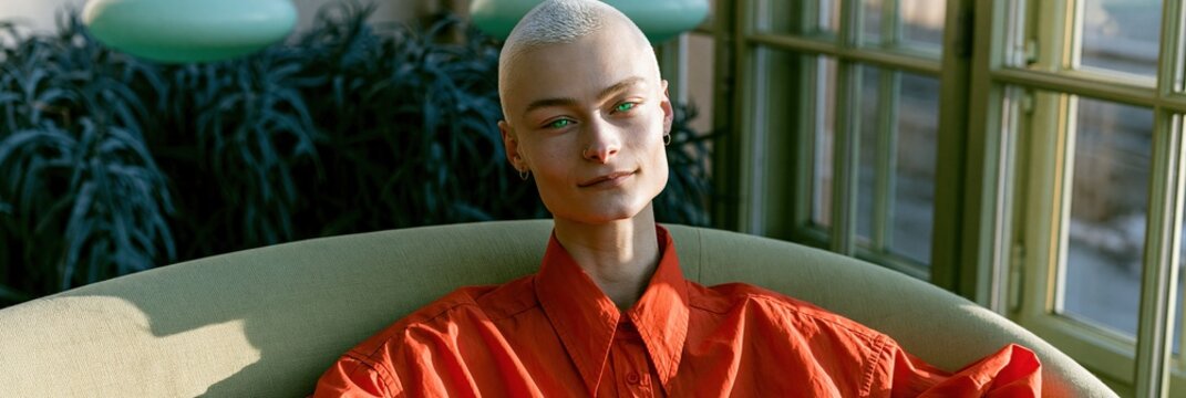 Panoramic close portrait of a bald model wearing a vivid red shirt on a curved sofa by a window with soft sunlight for editorial banner use